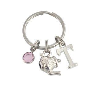 May include: Silver-toned keychain with a round key ring. Charms include a pink gemstone, a teapot, and the letter "T". The teapot charm has detailed floral designs. The keychain is designed for personal use.