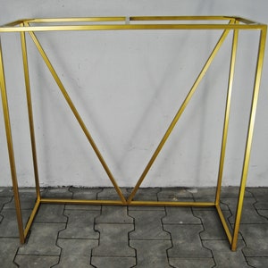 Op de afbeelding: Een gouden metalen standaard met een rechthoekig blad en een geometrisch ontwerp. De standaard is gemaakt van dunne metalen staven en heeft een eenvoudige, moderne stijl.