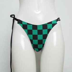 以下が含まれることがあります： 黒と緑のチェック柄のビキニボトム。黒い紐が付いています。ビキニボトムは、クラシックな三角形の形とローライズのウエストです。生地は正方形のパターンを繰り返しています。