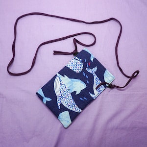 May include: A blue and white fabric pouch with a whale design and a brown adjustable strap. The pouch is perfect for carrying small items like a phone or keys.