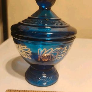May include: A deep blue glass candy dish with a matching lid, featuring gold-coloured floral patterns around the bowl. A wooden ruler is positioned at the base, showing the dish stands approximately 11.4 cm tall from the base to the lid top.