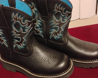 May include: A pair of black boots with a textured finish, featuring bright blue interior lining and intricate blue stitching patterns on the shaft. The boots have a rounded toe, pull tabs at the top, and a thick, dark gray lugged sole with visible stitching.