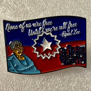 May include: A blue, red, and white enamel pin with a quote from Opal Lee: "None of us are free until we're all free." The pin features a portrait of Opal Lee and a stylized star.