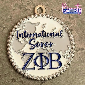 Puede incluir: Un colgante metálico circular con fondo blanco y un diseño de mapa mundial en gris claro. El texto International Soror ZΦB está escrito en letra cursiva y de imprenta azul oscuro. El borde está decorado con pequeñas cuentas plateadas.