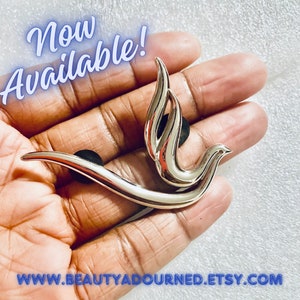May include: Silver-toned dove brooch with a stylized design. The dove is depicted in flight with its wings spread wide. The brooch is held in a hand, with the text "Now Available!" superimposed on the image.