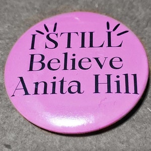 May include: Pink button with black text that reads "I STILL Believe Anita Hill".