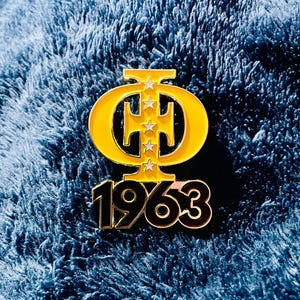 May include: A gold-colored pin with a yellow border featuring the Greek letters Phi and Omega, with three stars. The year "1963" is displayed in black and gold below the letters. The pin is set against a blue background.