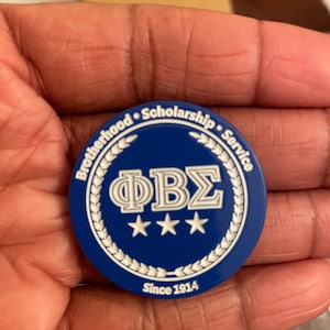 May include: A blue and white circular pin with the letters "ΦΒΣ" in white and three white stars below. The pin has a white wreath around the edge with the words "Brotherhood Scholarship Service" and "Since 1914" in white.
