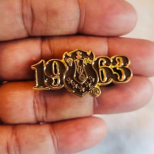 May include: A gold-colored pin with the numbers "1963" and a shield-shaped emblem. The emblem is brown with gold accents and features symbols and the Greek letters "ΦΘΕ". The pin has a small "R" symbol.