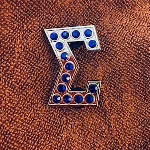 May include: A silver-colored letter 'E' with blue rhinestones. The letter is on a brown textured background.