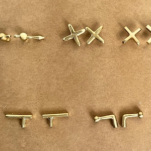 Peut inclure: Assortiment de quincaillerie de cabinet de couleur or, comprenant des boutons, des poignées et des tirettes de tiroir. La quincaillerie est disposée en rangées sur une surface brune.