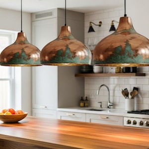 Peut inclure: Trois lampes suspendues en cuivre avec une finition en vert-de-gris sont suspendues au-dessus d'un îlot de cuisine en bois. Les lampes ont une forme de dôme et sont suspendues à des cordons noirs. La cuisine comprend des armoires blanches, une cuisinière en acier inoxydable et un bol de fruits.