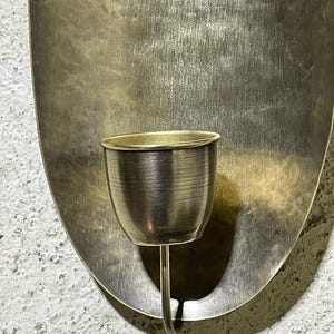 May include: A gold metal wall sconce with an oval-shaped backplate and a single candle holder. The candle holder is a small, round cup with a curved stem.