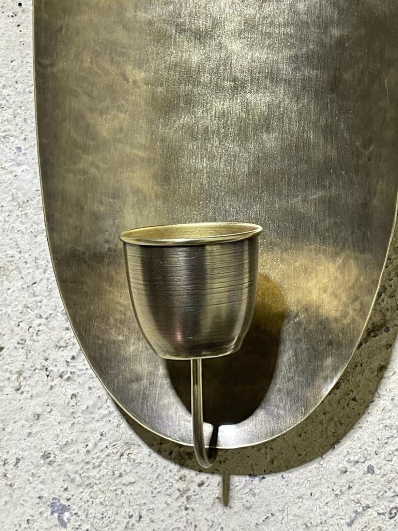 May include: A gold metal wall sconce with an oval-shaped backplate and a single candle holder. The candle holder is a small, round cup with a curved stem.