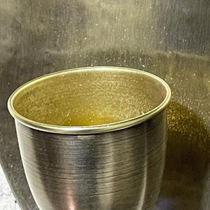 May include: A gold-colored metal cup with a smooth, shiny surface. The cup is empty and has a slightly rounded shape.