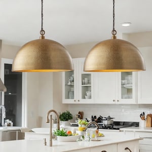 Puede incluir: Dos lámparas colgantes de cúpula de metal martillado dorado que cuelgan sobre una isla de cocina con gabinetes blancos y una encimera blanca.
