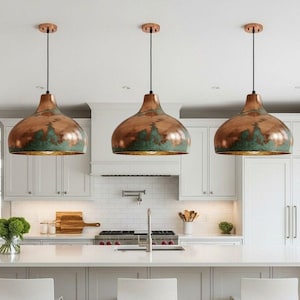 May include: Three copper pendant lights with a green patina and gold interior hang above a white kitchen island. The lights have a bell shape and are suspended by black cords. The kitchen features white cabinets and a white countertop.