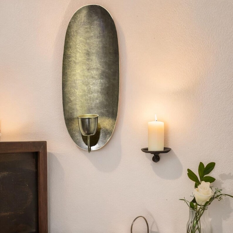 May include: A gold oval wall sconce with a candle holder. The sconce has a distressed finish and is mounted on a white wall. A second candle holder is shown on the wall below the sconce.