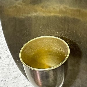 May include: A gold-colored metal wall sconce with a single light fixture. The sconce has a round, cup-shaped shade that is partially filled with a clear liquid.