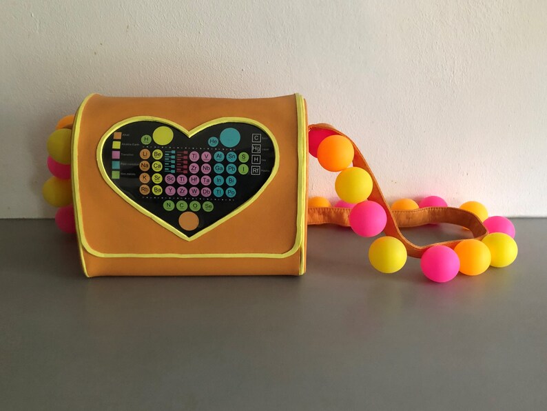 May include: A bright orange purse with a heart-shaped flap featuring a periodic table of elements. The purse has a bright yellow strap with a string of colorful balls.