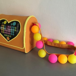 May include: A bright orange heart-shaped bag with a colorful pattern inside the heart. The bag has a yellow strap with a string of pink, yellow, and orange balls attached.