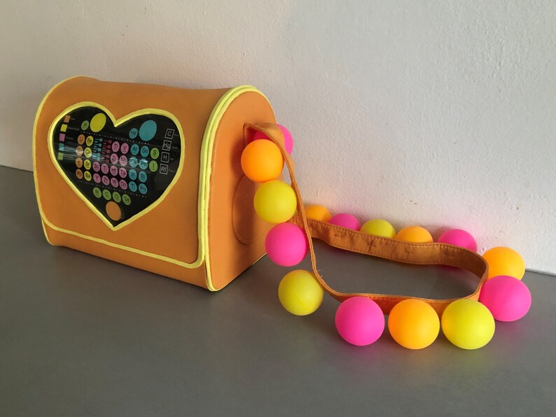 May include: A bright orange heart-shaped bag with a colorful pattern inside the heart. The bag has a yellow strap with a string of pink, yellow, and orange balls attached.
