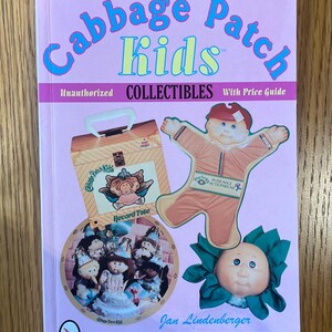 May include: A pink book titled "Cabbage Patch Kids" with the text "Unauthorized Collectibles With Price Guide." The cover features illustrations of Cabbage Patch Kids dolls and accessories. The author's name is Jan Lindenberger.