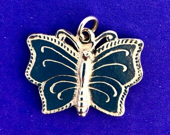 Puede incluir: Colgante de mariposa plateado con relleno de esmalte azul oscuro. La mariposa tiene patrones de alas detallados y un lazo en la parte superior para colgar. El colgante está sobre un fondo azul liso.
