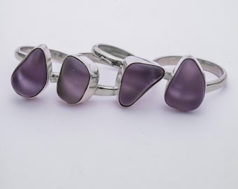 Puede incluir: Cuatro anillos de plata con engastes de piedras preciosas púrpuras. Los anillos están dispuestos en fila, con las piedras preciosas mirando al espectador.