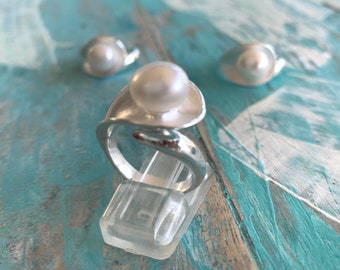 Puede incluir: Un anillo de plata con una perla blanca grande en el centro, exhibido en un soporte de plástico transparente. Dos pendientes de perlas a juego están ligeramente desenfocados en el fondo. La joyería está sobre una superficie pintada de azul y blanco.