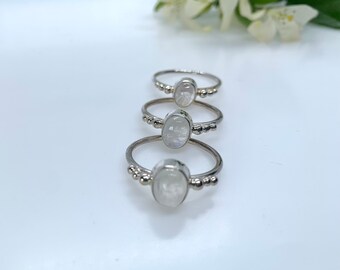 Puede incluir: Tres anillos de plata con cabujones ovalados de piedra lunar iridiscente están dispuestos en línea vertical. Cada anillo tiene una banda fina y cuentas decorativas de plata. Flores blancas y hojas verdes en el fondo.