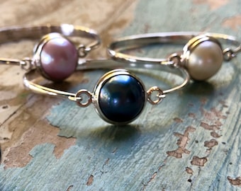 Puede incluir: Primer plano de tres pulseras de plata, cada una con una perla de diferente color: rosa, crema y azul oscuro. Las pulseras se exhiben sobre una superficie de madera azul claro desgastada.