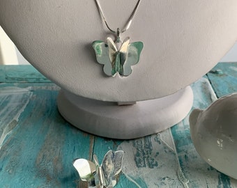 Puede incluir: Un collar con colgante de mariposa plateada y un anillo de mariposa a juego. El colgante y el anillo de mariposa tienen un diseño en capas con un toque de verde. La joyería se exhibe sobre un busto blanco contra una superficie de madera turquesa y blanca.