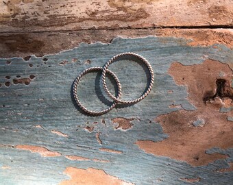 Puede incluir: Dos anillos plateados con diseño de cuerda trenzada. Los anillos son circulares y se encuentran sobre una superficie de madera envejecida y desgastada con tonos de pintura azul, marrón y blanca.