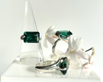 Puede incluir: Tres anillos de plata con grandes piedras preciosas rectangulares de color verde esmeralda. Un anillo se exhibe en un soporte triangular transparente. Flores blancas dispuestas alrededor de los anillos, sobre un fondo blanco.