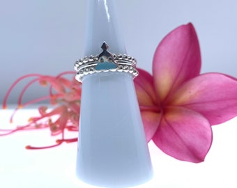 Puede incluir: Anillos de plata apilados con un diseño de Buda, exhibidos en un soporte cónico blanco. Los anillos tienen pequeños detalles de cuentas. Una flor rosa y capullos de flores rojas en el fondo.