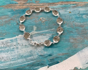 Puede incluir: Una pulsera de plata con una serie de eslabones texturizados en tono plateado. La pulsera está conectada por una delicada cadena y asegurada con un cierre de mosquetón. La pulsera se exhibe sobre una superficie de madera pintada de azul y desgastada.