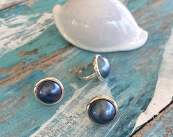 Puede incluir: Un conjunto de tres anillos de plata con piedras de color azul oscuro que parecen perlas. Los anillos están sobre una superficie de madera con una concha blanca.