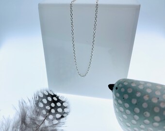 Puede incluir: Un delicado collar de cadena de plata con un diseño minimalista. El collar se muestra sobre un fondo blanco, con una pluma moteada y una figura de pájaro de cerámica en primer plano. La cadena mide aproximadamente 45 cm de largo.