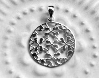 Puede incluir: Un colgante plateado con un diseño circular calado. El colgante presenta un intrincado patrón floral dentro del círculo y un pequeño lazo en la parte superior para una cadena. El fondo es una superficie blanca texturizada.