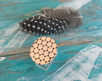 Puede incluir: Un anillo dorado con un diseño circular de pequeños círculos espaciados uniformemente. El anillo se exhibe sobre un soporte de plástico transparente, con una pluma estampada en el fondo. La pluma es negra y blanca con un patrón de lunares.