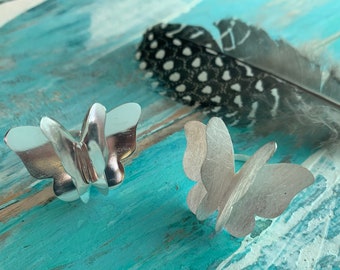 Puede incluir: Dos anillos de mariposa plateados con diseños de alas detallados sobre una superficie pintada en turquesa y azul. Una pluma moteada descansa cerca. Los anillos son una joya.