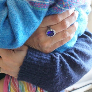 May include: A close-up of a hand wearing a ring with a large blue gemstone. The hand is holding a blue and pink striped jumper.