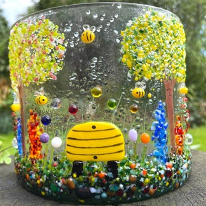 May include: A glass candle holder with a beehive design and a colorful mosaic of bees, flowers, and trees. The beehive is yellow with black stripes and is surrounded by a variety of colorful glass beads.