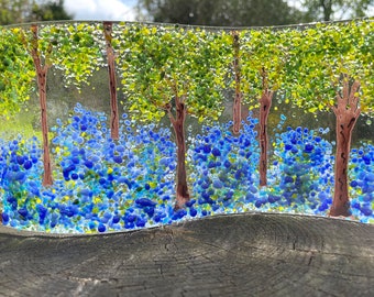 May include: A glass art piece depicting a forest scene with trees and blue flowers. The piece is made of glass and has a wavy shape.