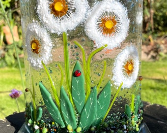 May include: A clear glass vase with a nature-inspired design. The vase features white dandelion flowers with yellow centers, green grass, and a ladybug. A yellow and black bee is also depicted. The vase sits on a clear glass base.