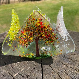 Könnte beinhalten: Eine dekorative Glasskulptur eines Baumes mit bunten Blättern in Rot-, Gelb- und Grüntönen. Der Baum ist in einer klaren, dreieckigen Glasstruktur mit gebogenen Kanten eingeschlossen. Die Skulptur steht auf einer verwitterten Holzoberfläche.