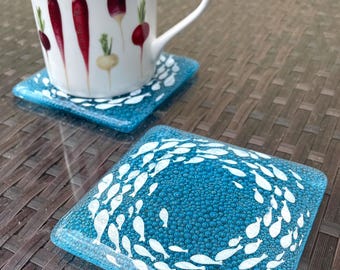 May include: Two blue glass coasters with white fish designs. The coaster on the left has a white background with a circle of fish. The coaster on the right has a white background with a circle of fish. The coasters are on a woven wicker surface.