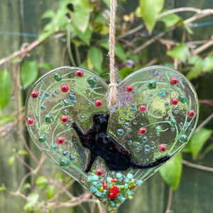 May include: A glass heart-shaped ornament featuring a black cat silhouette. The heart is decorated with green vines, red berries, and colourful glass beads.
