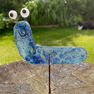 May include: A blue glass garden stake shaped like a smiling slug with large white eyes. The slug has a textured surface with small air bubbles.